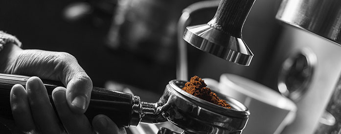 Imagen en blanco y negro, excepto el café molido que tiene su color natural, de un cacillo con dicho café molido sujetado por una mano para ser prensado y colocado en la máquina para preparar el mejor café espresso Jurado.