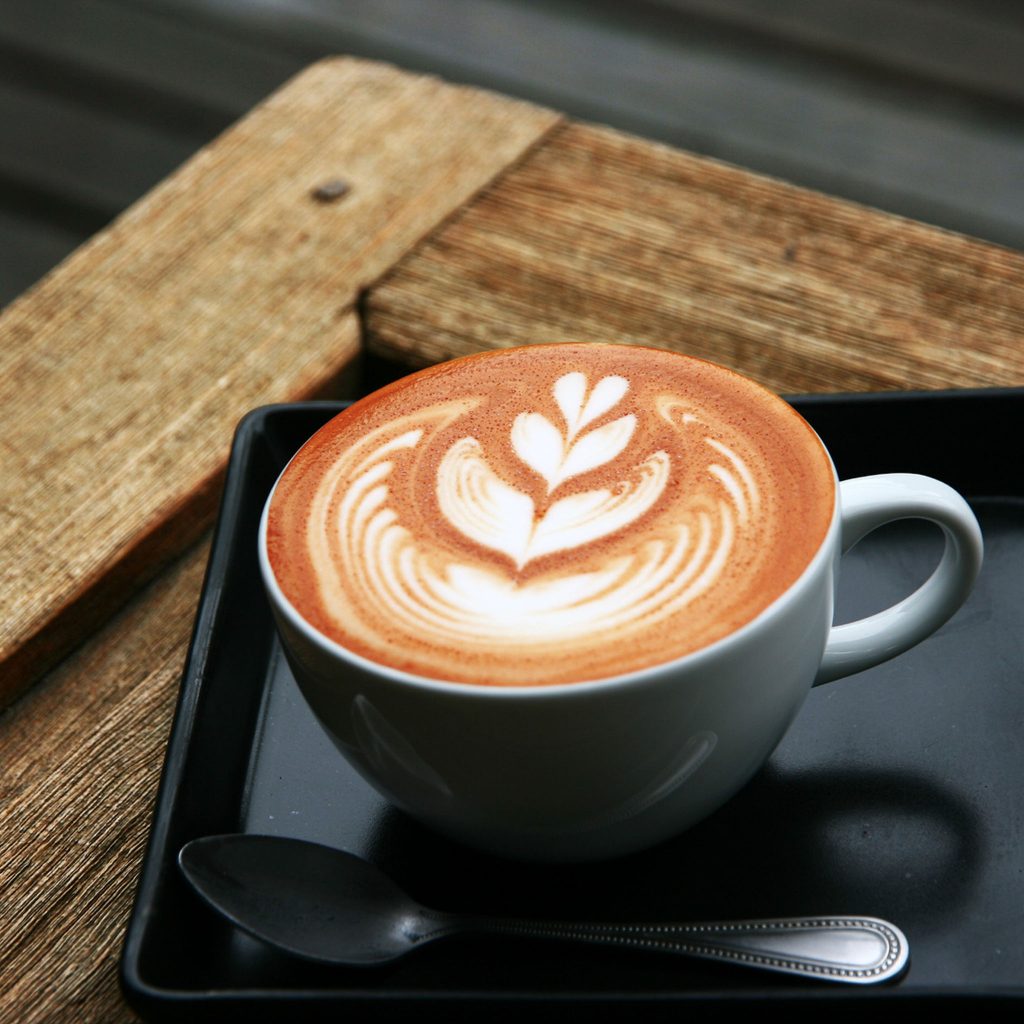 Imagen con una taza sobre un tablón de madera en el que vemos un ejemplo de Latte Art, mediante el método etching art con dibujo de tulipán.