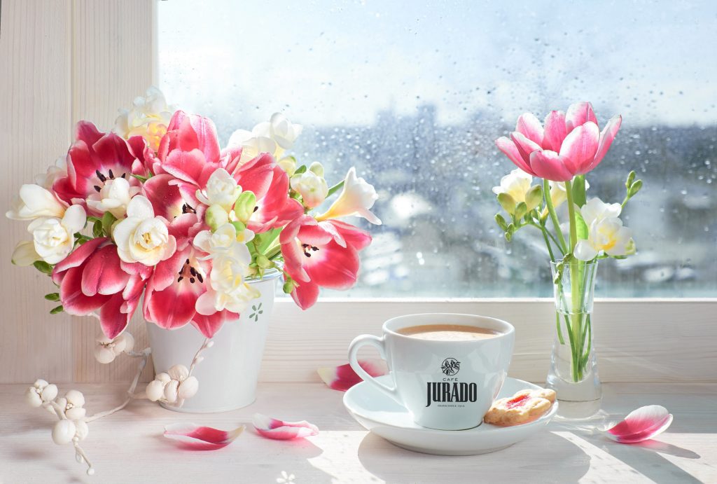 Imagen en la que se aprecia una taza de excelente café Jurado en el poyete de la ventana junto a un ramo de flores mientras entra los rayos de sol tras la lluvia de Primavera.