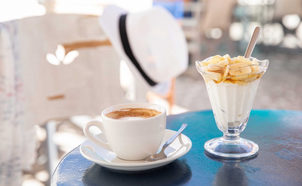 imagen veraniega en la que se ve un café recién hecho calentito junto a una copa con nata y plátano en un desayuno de verano.